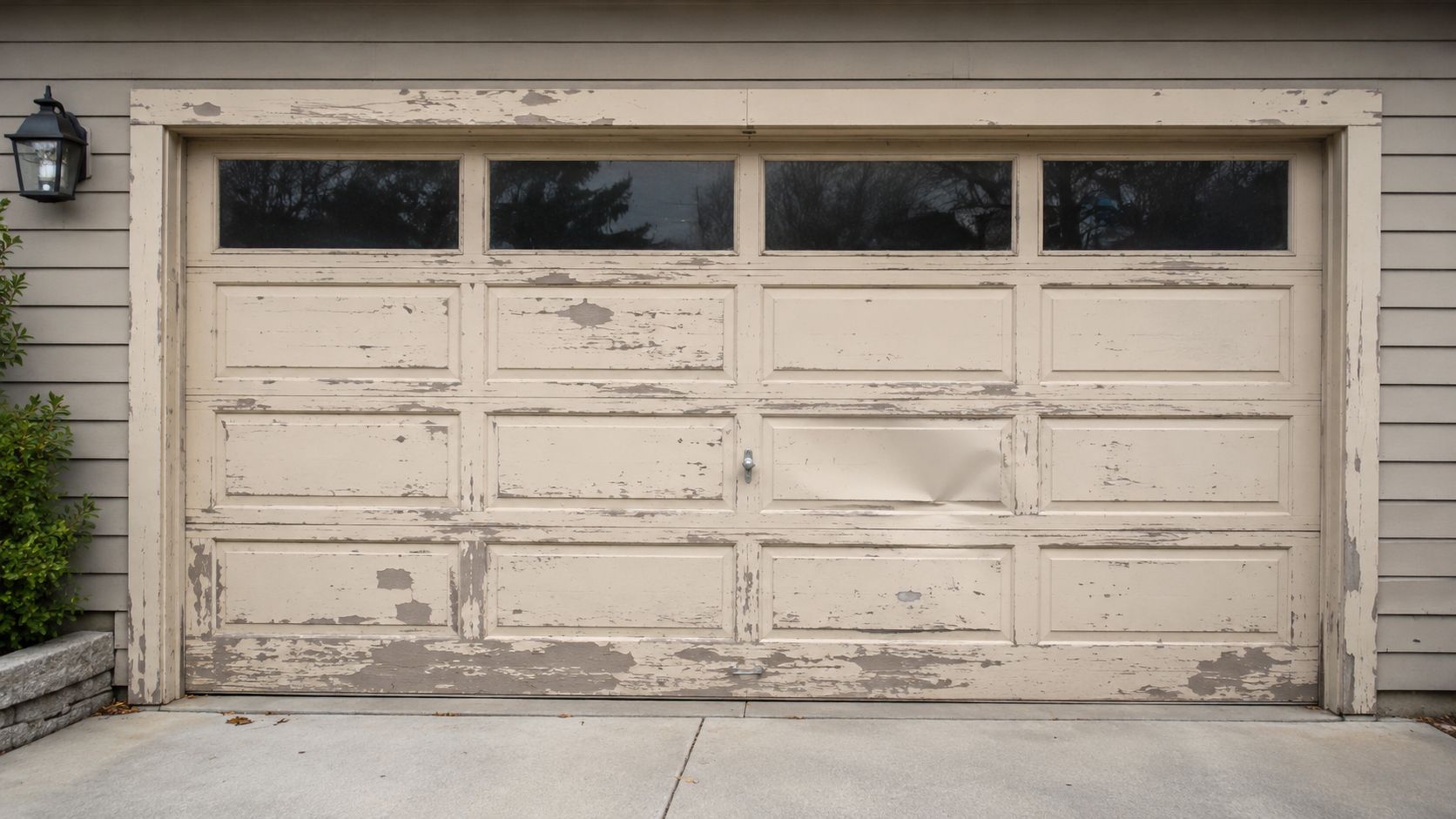A weathered, beige garage door with peeling paint, showing signs of wear, damage, and aging on a home.